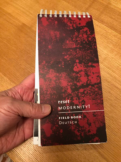 黒と赤のスパッタ模様の表紙を持つ小型スパイラルノート「reset MODERNITY! FIELD BOOK Deutsch」を手に持つ写真。